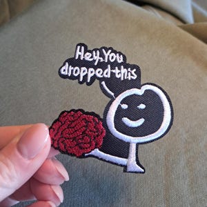 Op de afbeelding: Geborduurde patch met een zwart figuur, witte omtrek en een rood brein. De patch heeft de tekst "Hey, You dropped this" in het wit. De patch wordt getoond tegen een groene achtergrond.