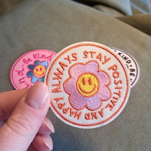 Könnte beinhalten: Drei gestickte Aufnäher mit positiven Botschaften. Der zentrale Aufnäher ist rund mit einer rosa Blume und einem gelben Smiley, umgeben von den Worten "ALWAYS STAY POSITIVE AND HAPPY". Die anderen Aufnäher sagen "Be Kind" und "Kind Be".