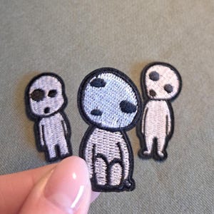 Könnte beinhalten: Drei gestickte Aufnäher mit einem cartoonartigen Waldgeist-Design. Die Aufnäher haben einen hellgrauen Körper mit schwarzen Akzenten und einem schwarzen Rand. Die Aufnäher befinden sich auf einem grünen Hintergrund.