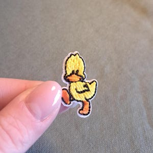 Op de afbeelding: Geborduurde patch van een gele eend met oranje voeten en snavel, omlijnd in zwart. Het eendje loopt, met één been omhoog. De patch heeft een witte rand en staat op een groene achtergrond.