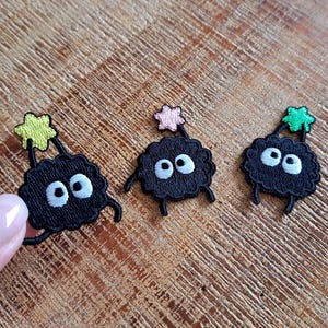 Può includere: Tre toppe ricamate nere raffiguranti sprite di fuliggine con occhi bianchi, piccole zampe nere e un accento a forma di stella in giallo, rosa e verde. Le toppe sono su una superficie di legno.