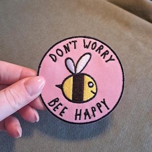 Può includere: Toppa rotonda ricamata rosa con un'ape dei cartoni animati e le parole "DON'T WORRY BEE HAPPY" in nero. L'ape è gialla e nera con ali bianche. La toppa è su uno sfondo verde.