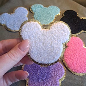 Könnte beinhalten: Eine Sammlung von Mickey Mouse-förmigen Aufnähern aus Plüsch in verschiedenen Farben. Die Aufnäher sind in Weiß, Blau, Grün, Schwarz, Rosa und Lila gehalten. Jeder Aufnäher hat einen goldglitzernden Rand.