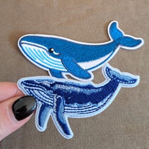 Könnte beinhalten: Zwei gestickte Wal-Aufnäher aus blauem und weißem Garn. Der obere Wal ist in mittlerem Blau mit weißem Bauch gehalten, während der untere Wal ein dunkelblaues, strukturiertes Muster mit hellblauem Rand und weißen Akzenten aufweist.