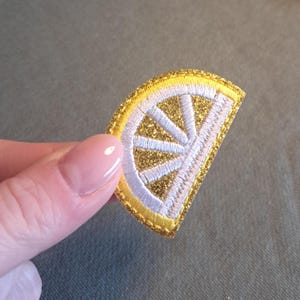 Könnte beinhalten: Gestickter Aufnäher in Form einer Zitronenscheibe. Der Aufnäher hat eine goldglitzernde Mitte, weiße und hellgelbe Details und einen goldenen Rand. Der Aufnäher wird vor einem grauen Hintergrund gehalten.