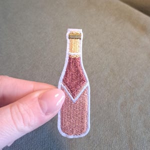 Può includere: Toppa ricamata a forma di bottiglia di vino. La toppa ha un bordo bianco, un collo marrone chiaro e un corpo rosa con un design a chevron bianco. La toppa è tenuta in mano.
