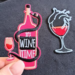 Può includere: Due toppe ricamate: una presenta una bottiglia di vino rosa con un serpente nero e la scritta "IT'S WINE TIME", e un calice. L'altra toppa è un calice con un disegno a forma di cuore riempito di liquido rosso.