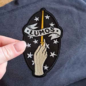 Könnte beinhalten: Schwarzer, gestickter Aufnäher mit einem Zauberstab, der von einer Hand gehalten wird, umgeben von Sternen. Das Wort "LUMOS" ist auf einem Banner abgebildet. Der Aufnäher hat eine ovale Form mit schwarzem Rand.