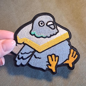 Könnte beinhalten: Gestickter Aufnäher mit einem Cartoon-Tauben-Design in Grau, Blau und Gelb. Der Aufnäher hat einen schwarzen Rand und zeigt den Vogel mit gelbem Kragen und orangefarbenen Füßen. Das Design ist detailliert und skurril.