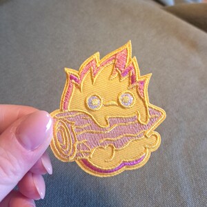 Könnte beinhalten: Gestickter Aufnäher mit einer cartoonartigen, flammenartigen Kreatur in Gelb und Rosa. Das Design umfasst ein stilisiertes Gesicht mit zwei runden Augen und einem Mund, der eine baumstammartige Form hält. Der Aufnäher hat ein detailliertes, gesticktes Aussehen.