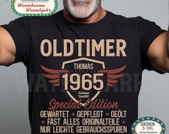 Può includere: T-shirt nera con la scritta "OLDTIMER" in lettere beige. Sotto, il nome "THOMAS" è sopra l'anno "1965" in un disegno a scudo con ali rosse. La maglietta presenta anche testo in tedesco, tra cui "Special Edition".