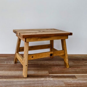 May include: A small, square wooden stool with a natural finish. The top is made of various wood pieces, and the legs are angled for stability. The stool is approximately 30 cm tall and suitable for various uses.