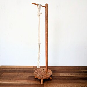 May include: A wooden plant stand with a round base and a single upright post. A white macrame plant hanger is hanging from the post.