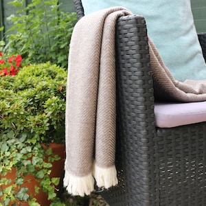 May include: A taupe throw blanket featuring a woven herringbone pattern and white fringe trim at the bottom edge. The blanket is draped over the arm of a black woven chair, with a light blue cushion visible behind the fabric.