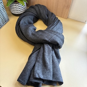 Könnte beinhalten: Ein dunkelgrauer Schal aus weichem Strickmaterial. Der Schal ist drapiert und gefaltet, wodurch seine Länge und Textur hervorgehoben werden. Der Schal ist einfarbig und für verschiedene Outfits geeignet.