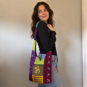 May include: A hobo-style shoulder bag featuring a patchwork design in purple, lime green, and teal. The bag includes a central olive green pocket with a gold Om symbol, accented by side panels with decorative white cross-stitching and a button closure.