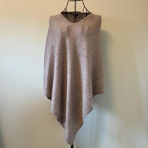 May include: A light brown poncho with a V-neck design. The poncho is made of a soft, textured material and drapes over the shoulders. The garment is displayed on a black metal mannequin.