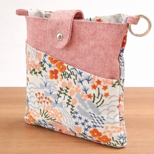 Puede incluir: Peque&ntilde;o estuche de fieltro rosa con un bolsillo estampado floral. El estuche tiene un cierre de solapa con un broche plateado y un llavero dorado. El estampado floral incluye flores y follaje naranja, azul y blanco.