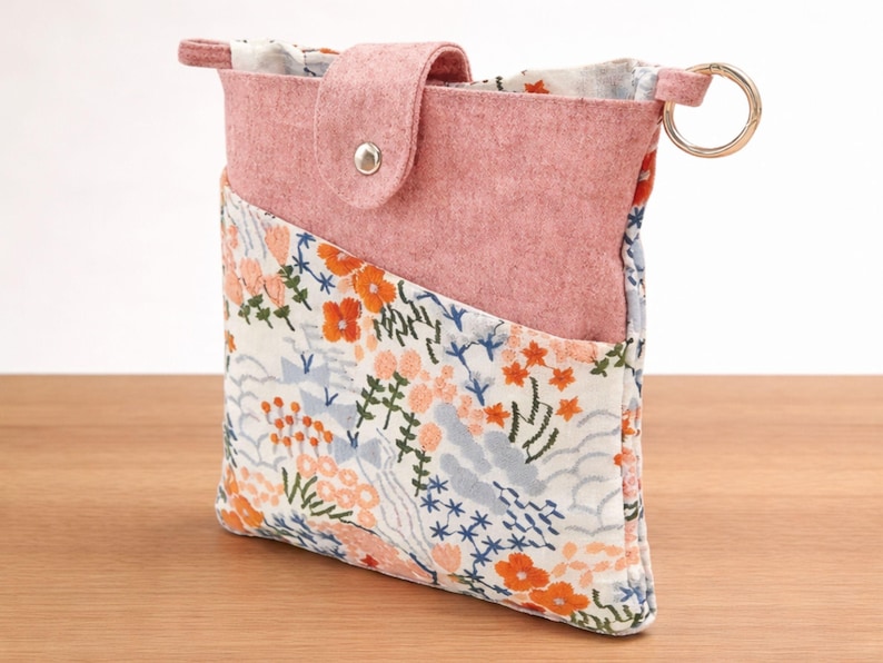 Puede incluir: Peque&ntilde;o estuche de fieltro rosa con un bolsillo estampado floral. El estuche tiene un cierre de solapa con un broche plateado y un llavero dorado. El estampado floral incluye flores y follaje naranja, azul y blanco.