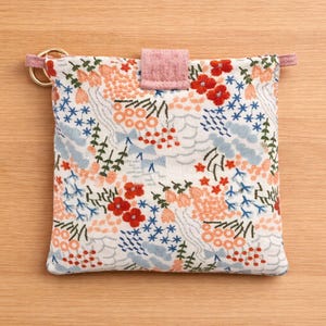 Puede incluir: Una bolsa cuadrada con estampado floral, con cierre de solapa rosa y detalles de anillo dorado. La tela presenta un fondo blanco con flores y follaje bordados en rojo, azul y melocot&oacute;n. Accesorio decorativo.