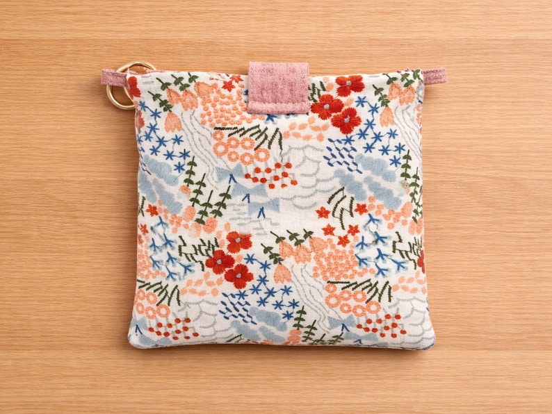 Puede incluir: Una bolsa cuadrada con estampado floral, con cierre de solapa rosa y detalles de anillo dorado. La tela presenta un fondo blanco con flores y follaje bordados en rojo, azul y melocot&oacute;n. Accesorio decorativo.