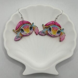May include: A pair of dangle earrings featuring a colourful cartoon turtle design. The turtles have pink shells, blue and yellow accents, and large, bright eyes.