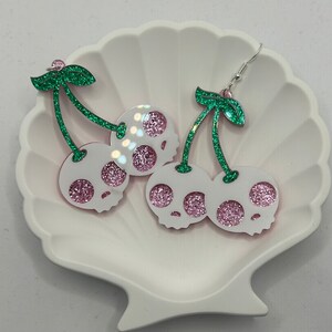May include: A pair of dangle earrings featuring a pink and green glitter skull cherry design.