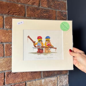Op de afbeelding: Waterverfschilderij van twee Lego-figuren gekleed als zeelieden, een in rood en de ander in blauw. Ze houden houten gereedschap vast. De schildering is op crèmekleurig papier en ingelijst.