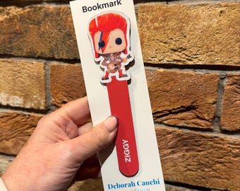 May include: Red and white bookmark with a pop culture figure of David Bowie as Ziggy Stardust. The bookmark has the text "Bookmark" at the top, "Ziggy" at the bottom, and "Deborah Cauchi toysfart.co.uk" at the bottom.