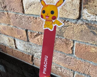 Peut inclure: Un marque-page rouge avec une illustration du personnage jaune Pikachu et le mot "PIKACHU" imprimé dessus.