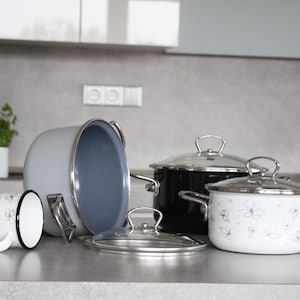 Op de afbeelding: Een set van vier potten met deksels, twee met witte bloemenmotieven, een zwart en een grijs. De potten staan op een grijs aanrecht.