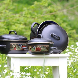 May include: Four black enamel pots with floral designs. The pots are arranged on a white wooden stool. The pots are of various sizes and have a gold rim.