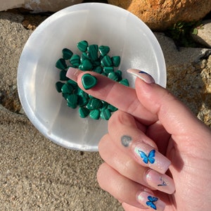 May include: A white bowl filled with heart-shaped malachite stones. A single malachite heart rests on a finger. The background includes rocks and a stone surface. The nails have butterfly designs.