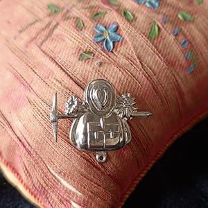 Può includere: Una spilla in metallo color argento raffigurante una figura centrale con cappello, affiancata da un piccone a sinistra e un fiore a destra. La figura presenta un dettaglio a forma di borsa rettangolare alla base e un piccolo occhiello circolare.