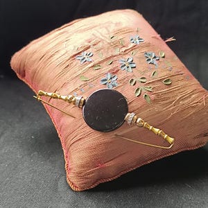 Puede incluir: Un broche de metal dorado con un disco negro circular central, flanqueado por dos pequeños anillos plateados con piedras claras y segmentos metálicos con textura de bambú, sobre un pequeño cojín cuadrado de tela color melocotón con bordados.