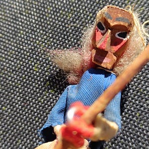 Peut inclure: Une marionnette en bois faite à la main avec un visage peint, des cheveux blonds et une chemise bleue. La marionnette tient un bâton en bois avec un objet rouge attaché. La marionnette est posée sur un fond gris foncé texturé.