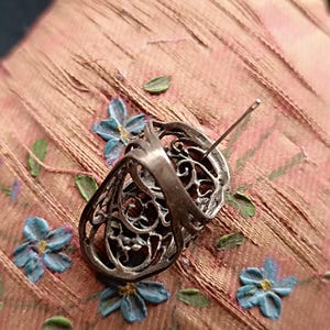 Peut inclure: Une boucle d'oreille ovale en argent présentant un design complexe en filigrane ajouré avec des motifs métalliques tourbillonnants. La boucle d'oreille comprend une tige fine et droite à l'arrière, mettant en valeur une structure en métal détaillée.