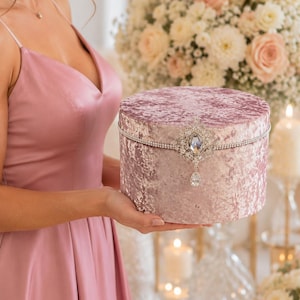 May include: A round, dusty rose crushed velvet box featuring a silver-toned rhinestone trim around the lid. The center of the lid is adorned with a large, ornate silver brooch set with clear crystals and a dangling teardrop-shaped crystal pendant.