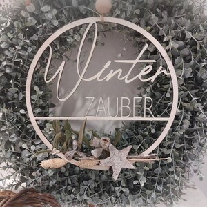 Pu&ograve; includere: Una ghirlanda di legno bianco con le parole "Winterzauber" scritte su di essa. La ghirlanda &egrave; decorata con foglie di eucalipto e una stella argentata.