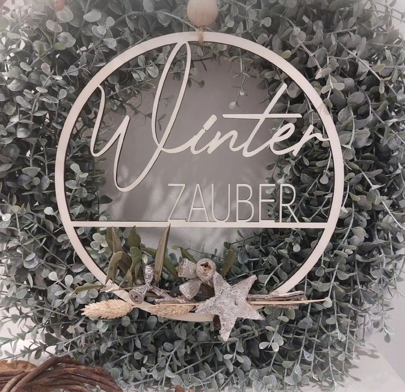 Pu&ograve; includere: Una ghirlanda di legno bianco con le parole "Winterzauber" scritte su di essa. La ghirlanda &egrave; decorata con foglie di eucalipto e una stella argentata.