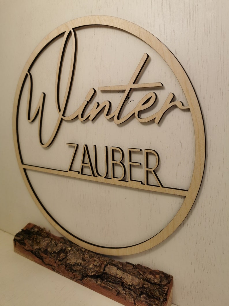 Pu&ograve; includere: Un cerchio di legno con la scritta "Winterzauber" su di esso. Il cerchio &egrave; su una base di legno.