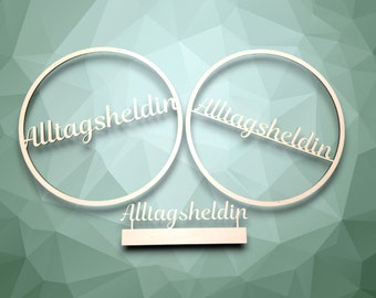 May include: Two wooden circles with the text "Alltagsheldin" written on them. The circles are connected by a smaller wooden rectangle with the same text written on it.