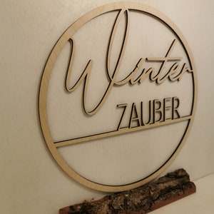 Pu&ograve; includere: Un cerchio di legno con la parola "Winterzauber" scritta in corsivo. Il cerchio &egrave; montato su un piccolo tronco di legno dall'aspetto naturale.