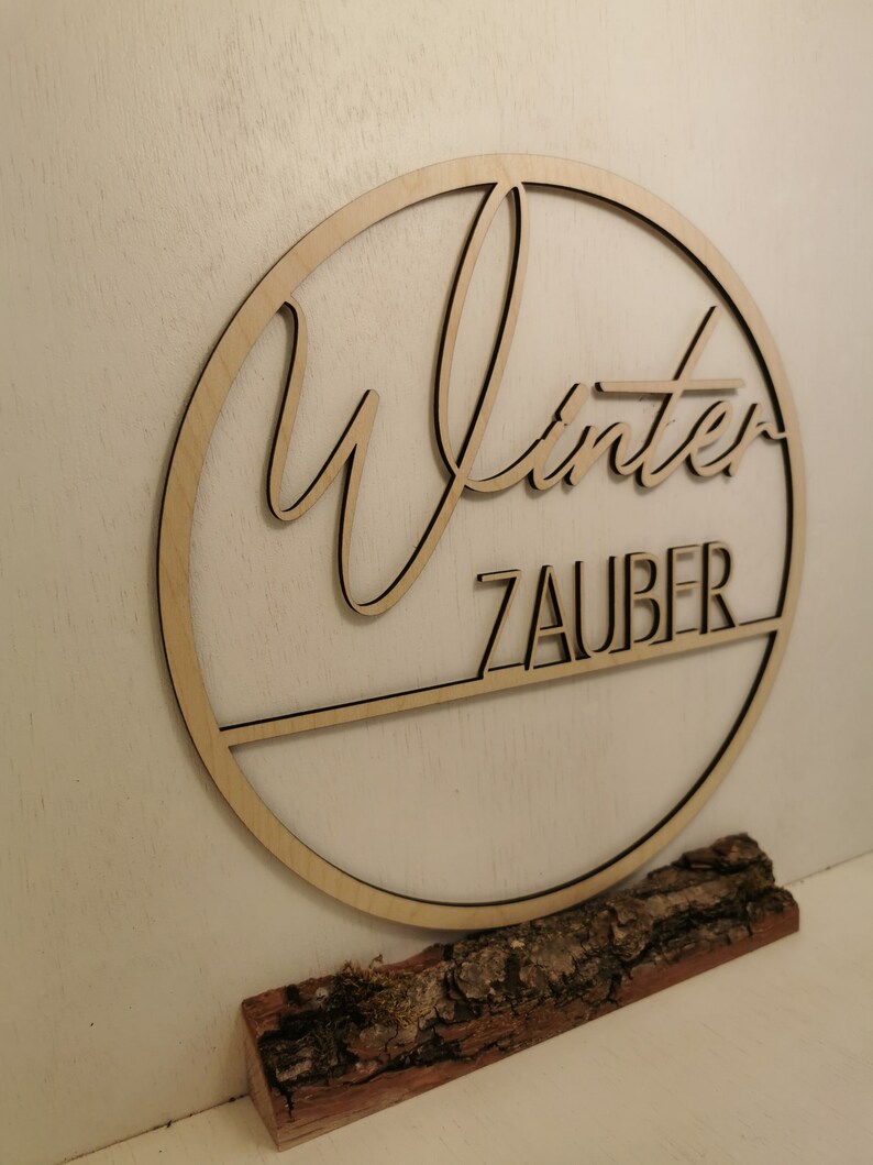 Pu&ograve; includere: Un cerchio di legno con la parola "Winterzauber" scritta in corsivo. Il cerchio &egrave; montato su un piccolo tronco di legno dall'aspetto naturale.