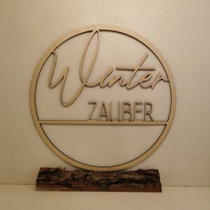 Pu&ograve; includere: Un cerchio di legno con la parola "Winterzauber" scritta in corsivo. Il cerchio &egrave; montato su un pezzo di corteccia d'albero.