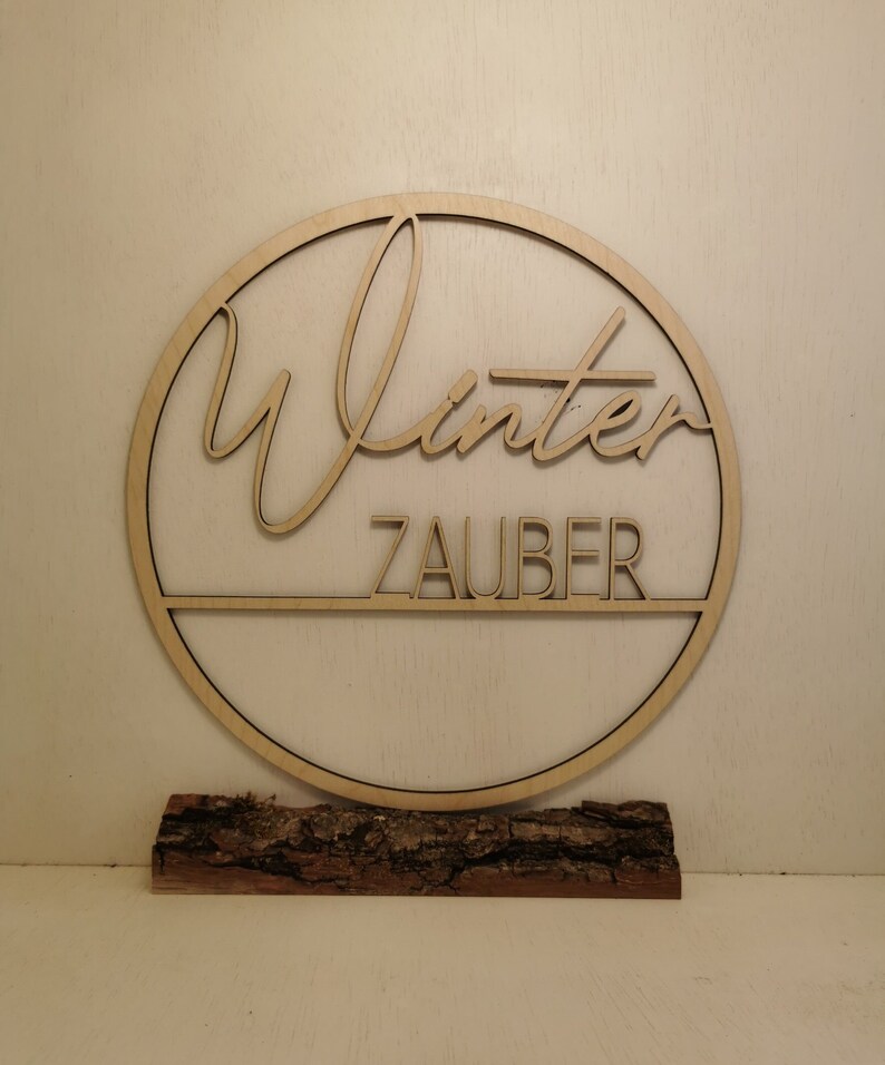 Pu&ograve; includere: Un cerchio di legno con la parola "Winterzauber" scritta in corsivo. Il cerchio &egrave; montato su un pezzo di corteccia d'albero.
