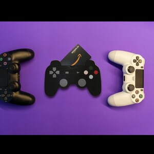 Könnte beinhalten: Zwei schwarze und weiße Videospiel-Controller mit einer Amazon-Geschenkkarte in der Mitte. Die Controller befinden sich auf einer violetten Oberfläche.