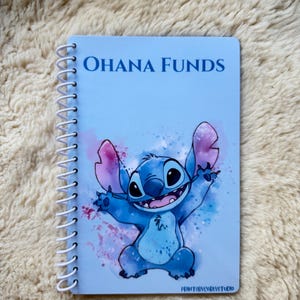 May include: A light blue spiral-bound notebook featuring a watercolour-style illustration of the character Stitch with arms raised. The text "OHANA FUNDS" is printed in dark blue at the top, and "PRINTERYCARLYSTUDIO" is printed at the bottom.
