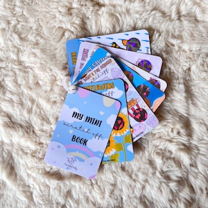 May include: A set of colourful scratch-off cards with various themes, including a duck, sunflowers, and a popcorn bucket. The cards are arranged in a fan shape and have a blue and pink colour scheme. The text on the front card reads "My Mini Scratch Off Book".