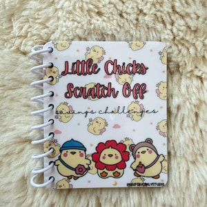May include: A spiral-bound notebook with a white cover featuring the text "Little Chicks Scratch Off Savings Challenges." The cover is decorated with cartoon chicks and illustrations. The notebook is designed for savings challenges.